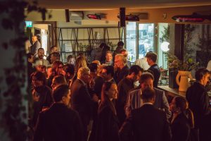 A lively indoor gathering unfolds as guests mingle and converse under warm ambient lighting. The space feels vibrant and refined, with layered textures, soft glows, and a dynamic crowd creating an engaging, social atmosphere.