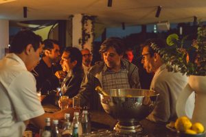 Guests gather around a warmly lit bar, engaged in relaxed conversation as drinks are prepared. The intimate lighting, layered textures, and lively crowd create a welcoming atmosphere that reflects a refined yet casual evening scene.