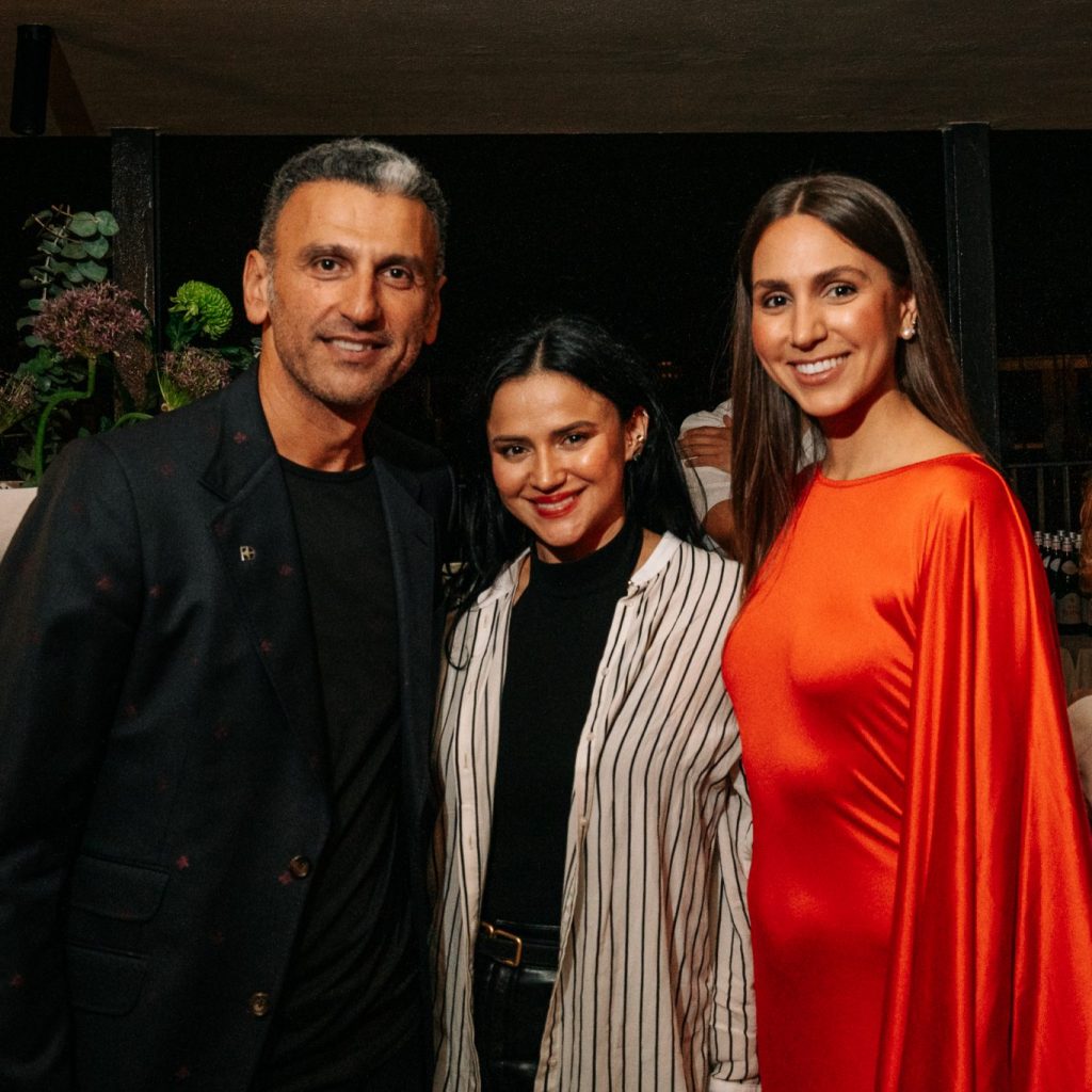 Three guests pose closely together, smiling against a softly lit evening backdrop. Elegant attire and bold color accents create a refined yet lively mood, capturing the essence of a stylish social gathering.