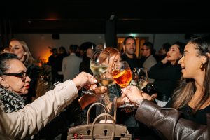 A lively toast brings guests together as glasses meet mid-air, capturing a spontaneous moment of celebration. Warm lighting and animated expressions convey the energy of a social evening filled with connection and shared enjoyment.