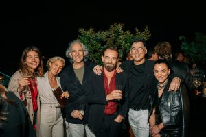 A group of guests gathers for a posed moment, smiling with drinks in hand under soft evening lighting. The mix of tailored looks and relaxed expressions captures a sociable, celebratory atmosphere in an elegant outdoor setting.