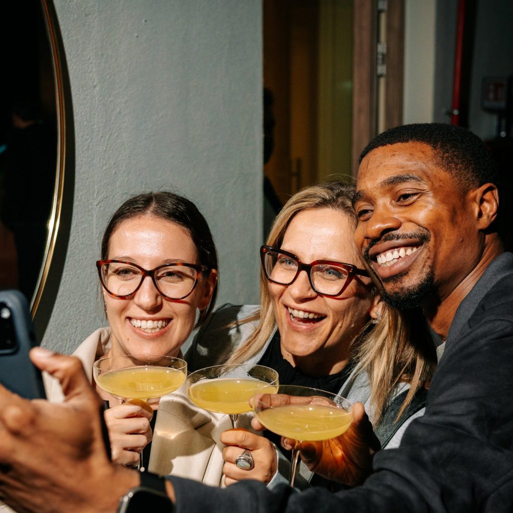 Three friends smile and lean in for a selfie, holding cocktails in a lively indoor setting. Warm lighting and close framing capture a joyful, intimate moment, highlighting connection, celebration, and a relaxed social atmosphere.
