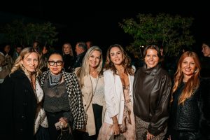 A group of elegantly dressed guests stands together at an evening event, smiling toward the camera. Soft lighting and natural greenery in the background create a warm, refined atmosphere with a relaxed, social energy.