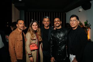 A well-dressed group gathers indoors, posing closely with relaxed smiles. Soft, warm lighting and dark surroundings enhance textures and tones, creating an intimate, stylish scene with a contemporary social ambiance.