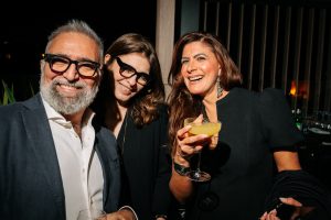 Three guests smile closely together, enjoying cocktails in a warm, dimly lit setting. The intimate framing and soft highlights emphasize expressions and elegance, capturing a lively, refined moment of social connection.