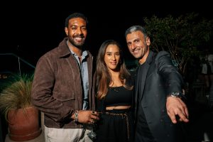 Three guests pose together at a nighttime gathering, smiling warmly toward the camera. Soft lighting highlights their expressions and textures, creating an intimate, polished scene with a relaxed yet sophisticated social vibe.