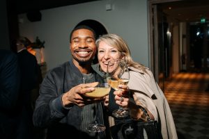 Two guests lean in with bright smiles, raising their drinks toward the camera in a cheerful toast. Warm lighting and close framing capture a lively, celebratory mood, highlighting a spontaneous moment of connection.