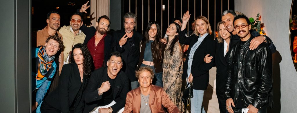 A large group gathers for an energetic photo, smiling, gesturing, and leaning in with playful expressions. The mix of styles and lively poses captures the dynamic spirit of a celebratory evening among friends.