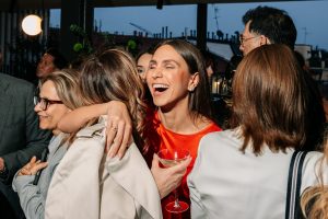 A joyful embrace unfolds amid a lively crowd, as guests share laughter and conversation. Soft evening light and candid expressions highlight the warmth and spontaneity of a vibrant social gathering.